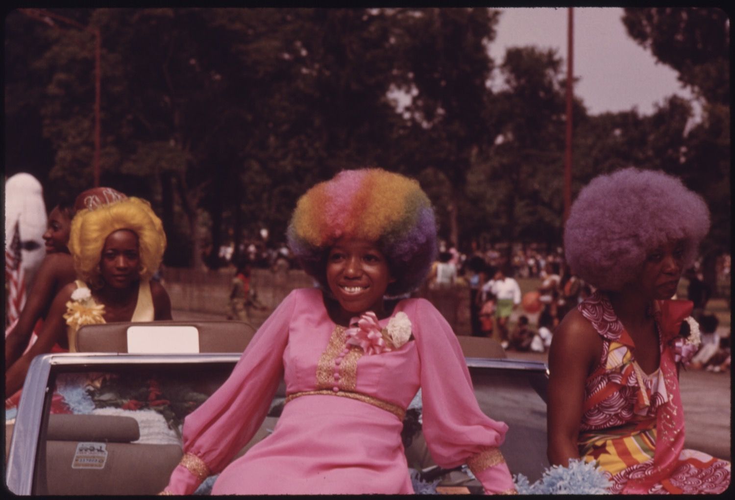 Three African American women with colorful afros ride in a convertible car during a parade or celebration.