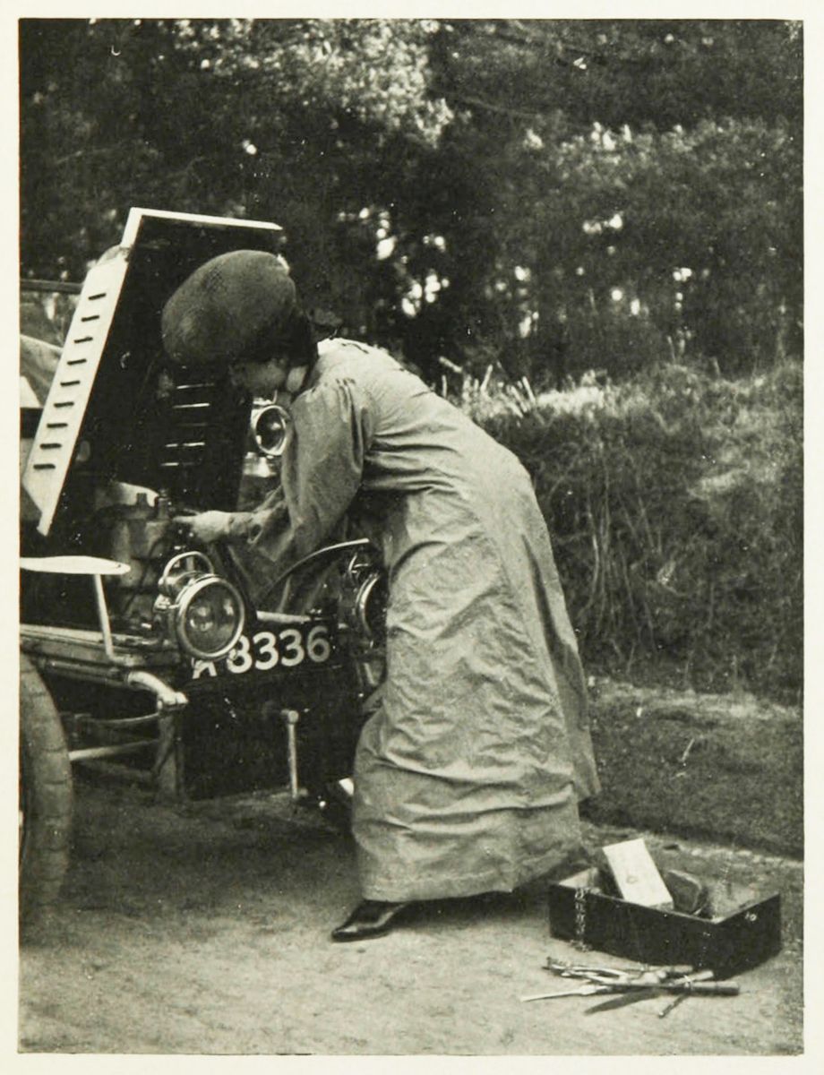A vintage black and white photograph of a woman working on the engine of a car in a rural setting.