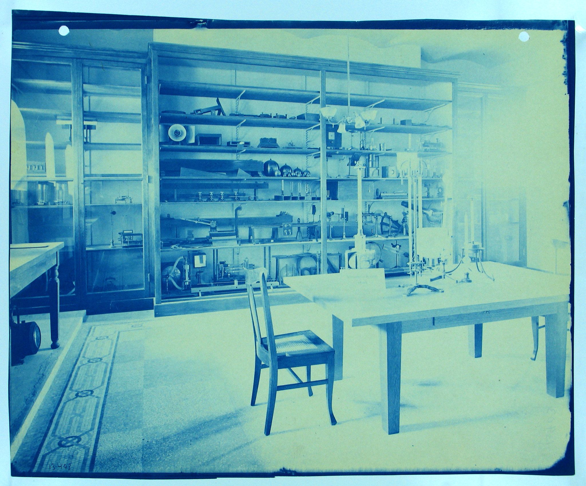 A cyanotype photograph of a room filled with shelves of objects, a table, and a chair.