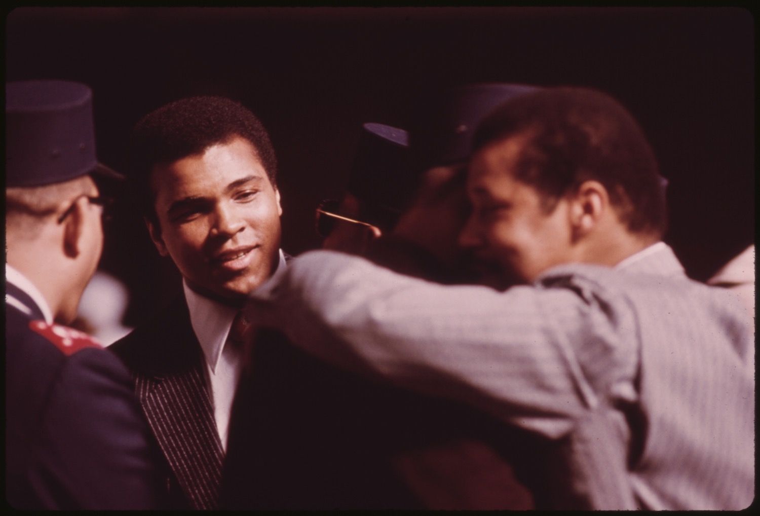 Close-up of Muhammad Ali in a suit, being embraced by another man, with a man in a hat to his left.