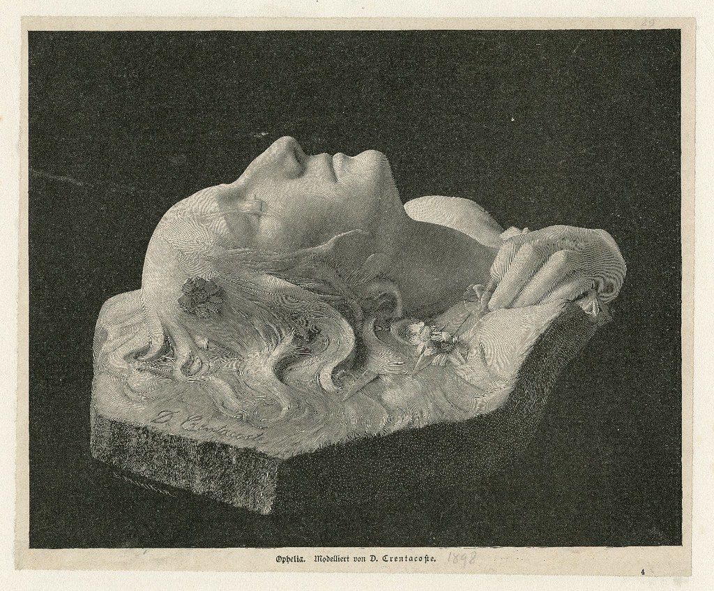 A black and white photograph of a sculpture depicting a woman, likely Ophelia from Shakespeare's Hamlet, as indicated by the text at the bottom of the image. The sculpture is a bust, showing the woman's head and upper torso. Her face is serene and tilted upwards, with closed eyes, suggesting a state of peacefulness or death. Her hair is long and wavy, cascading around her shoulders and blending with the flowing drapery of her clothing. The drapery is intricately detailed, with numerous folds and curves that create a sense of movement and texture. The sculpture is rendered in a realistic style, with attention to anatomical detail and the play of light and shadow. The background is dark and indistinct, which helps to emphasize the sculpture as the focal point of the image. The photograph is a reproduction of a sculpture by D. Cruentake, as indicated by the text at the bottom of the image, and is dated 1884.