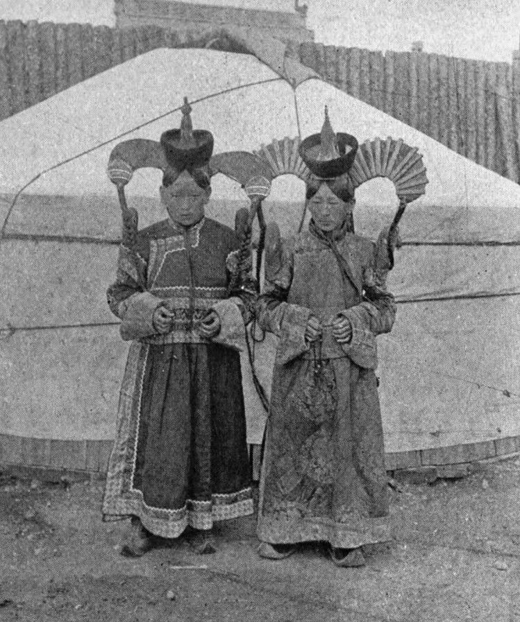 Two young people in traditional clothing stand in front of a yurt, each holding a fan-like object.
