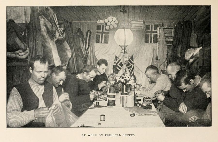 A black and white photograph of a group of men working around a long table in a rustic interior, focused on sewing or repairing clothing.