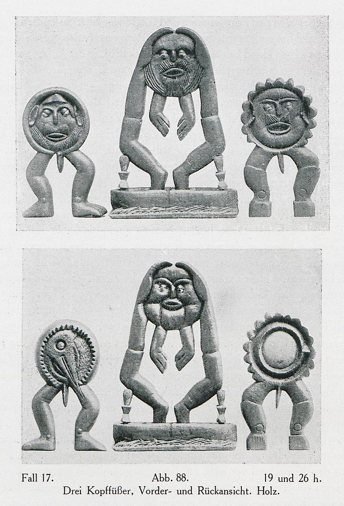 Black and white photograph of six wooden sculptures arranged in two rows of three. The sculptures are stylized human figures with elongated bodies and large heads, carved with expressive faces and minimal detail. They are mounted on rectangular bases.