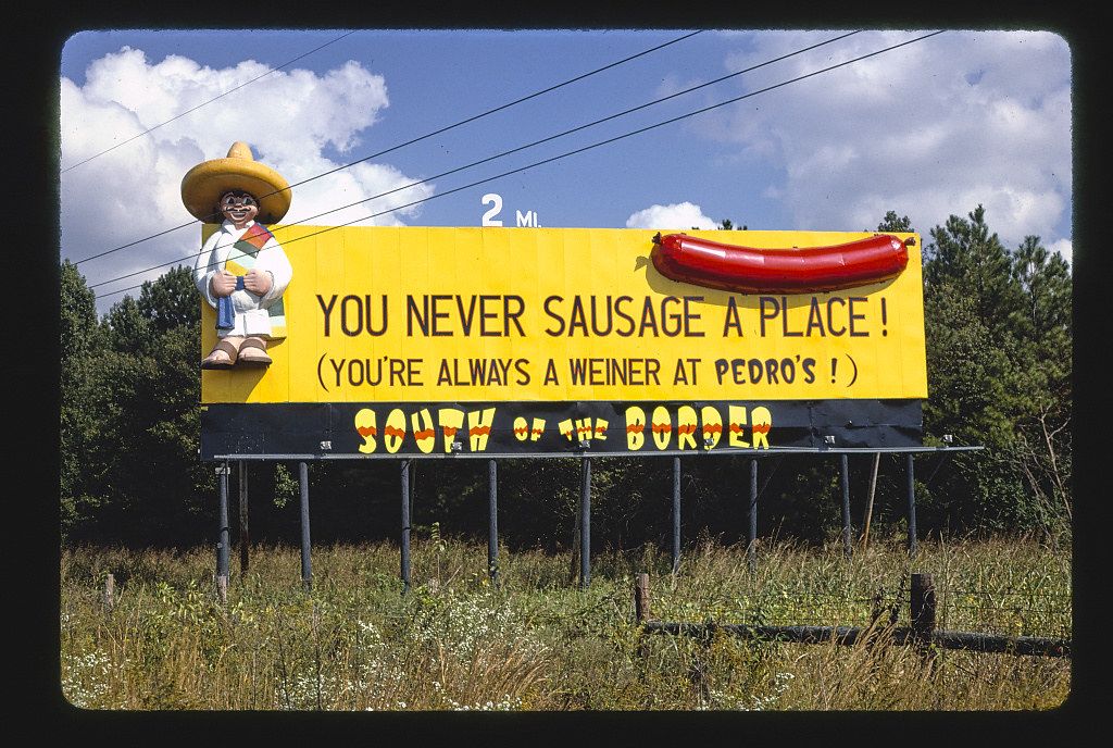 A vintage-style photograph of a large, colorful billboard advertising "South of the Border," featuring a cartoon man in a sombrero and a giant hot dog, set against a backdrop of a blue sky and green trees.