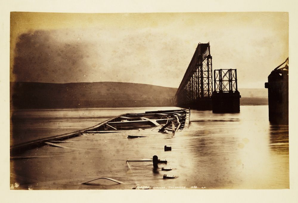 Sepia-toned historical photograph of a collapsed railway bridge in a wide body of water, with a hilly landscape and cloudy sky in the background.
