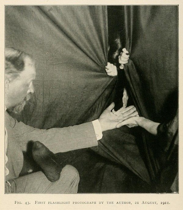 A vintage black and white photograph labeled 'First Flashlight Photograph by the Author, 21 August, 1911'. It depicts a person partially visible on the left and a pair of hands emerging from behind a dark, draped fabric.