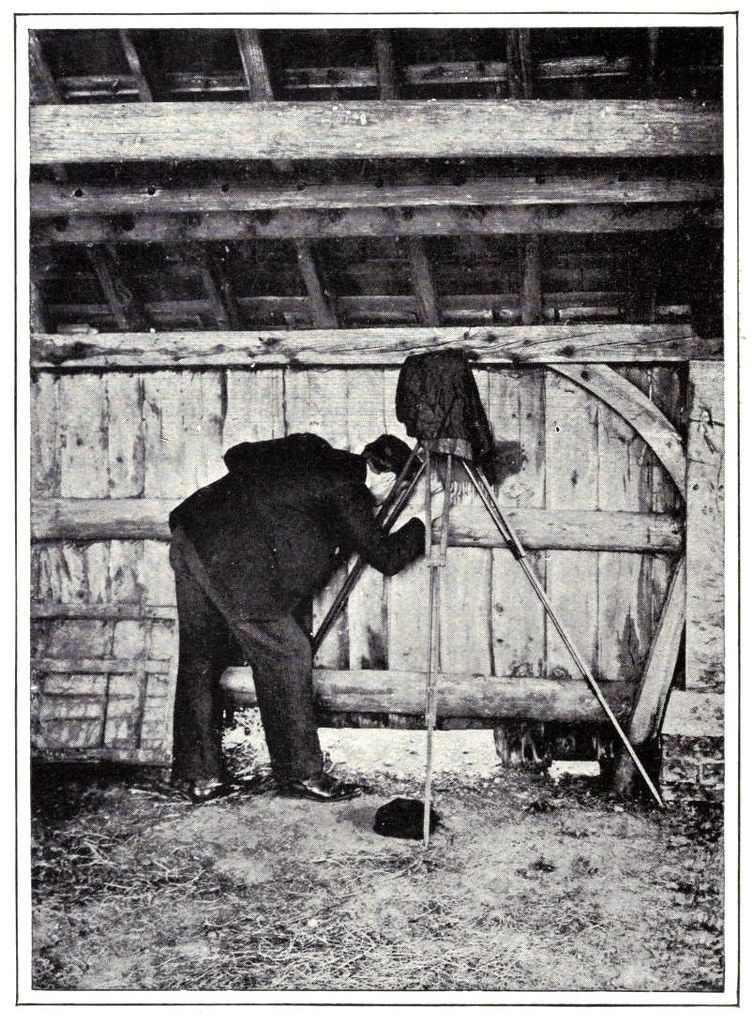 A black and white photograph of a photographer operating a large format camera on a tripod inside a rustic barn or shed.
