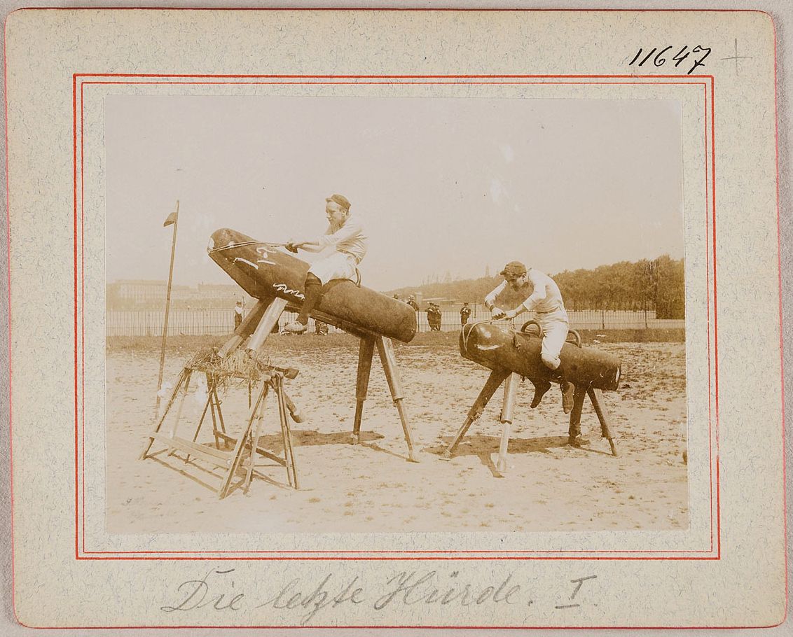 Vintage photograph of two men on horse-like wooden structures with cigar-shaped gliders, labeled 'Die luppe Meinde, I'.
