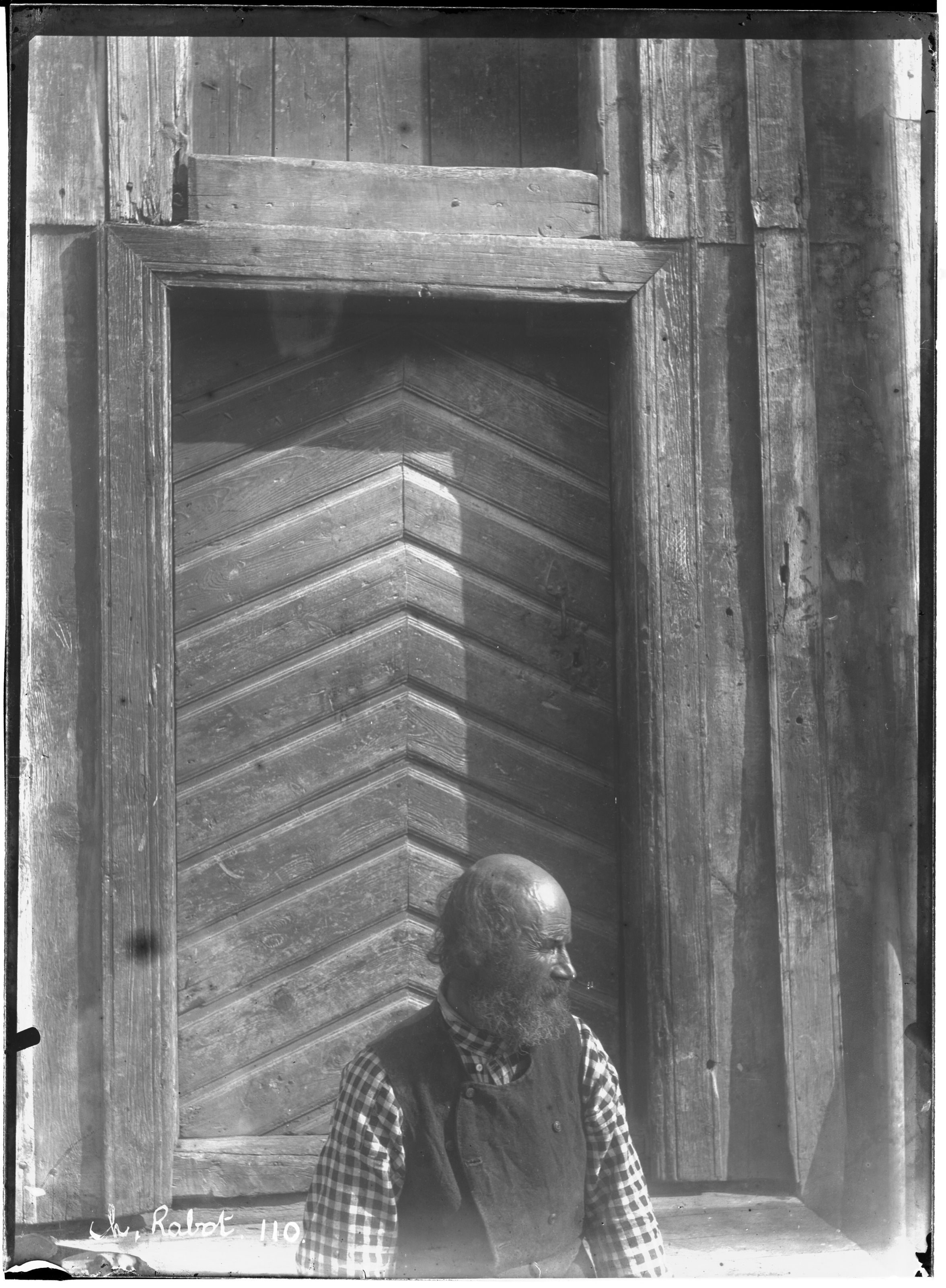 A black and white photograph of an elderly man seated in a rustic wooden doorway.