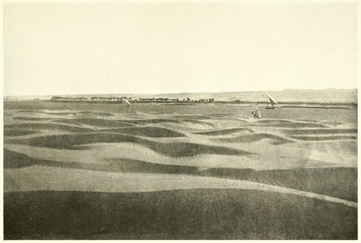 Vintage sepia-toned photograph of a desert landscape with sand dunes and boats on a body of water.