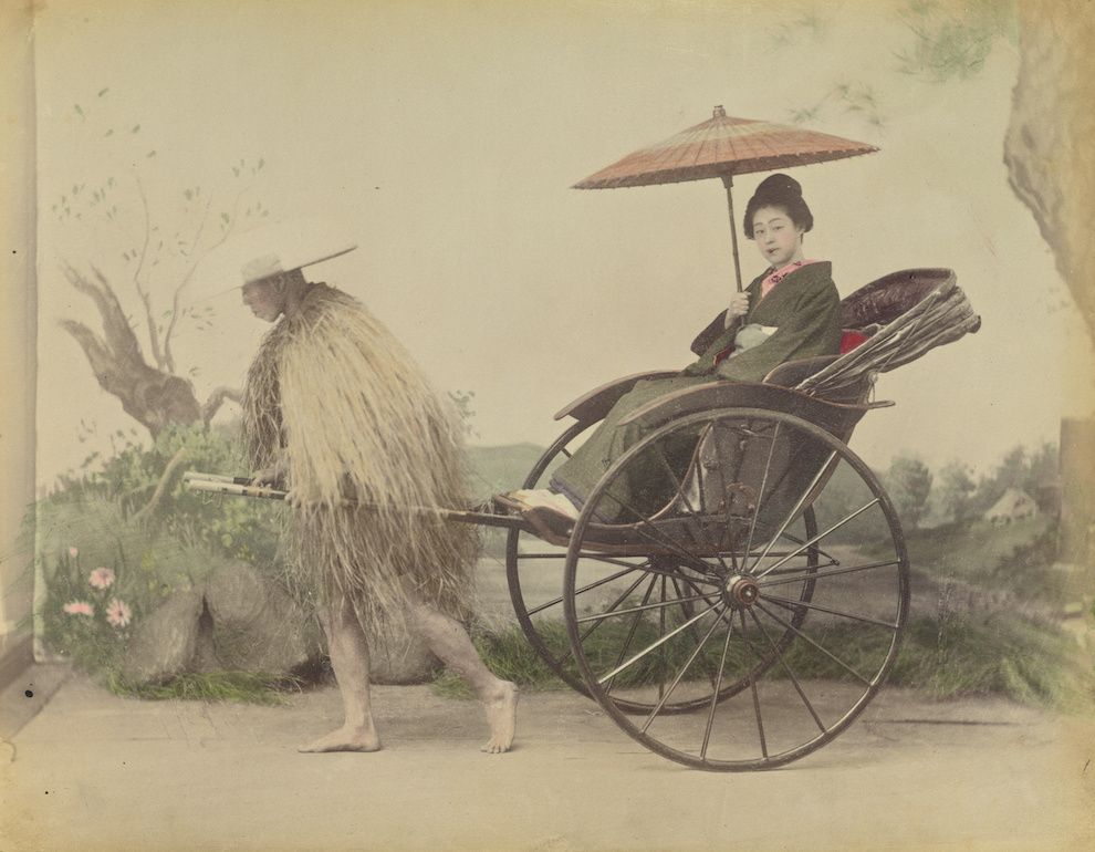 A vintage photograph of a woman in a kimono being pulled in a rickshaw by a man in a straw raincoat.