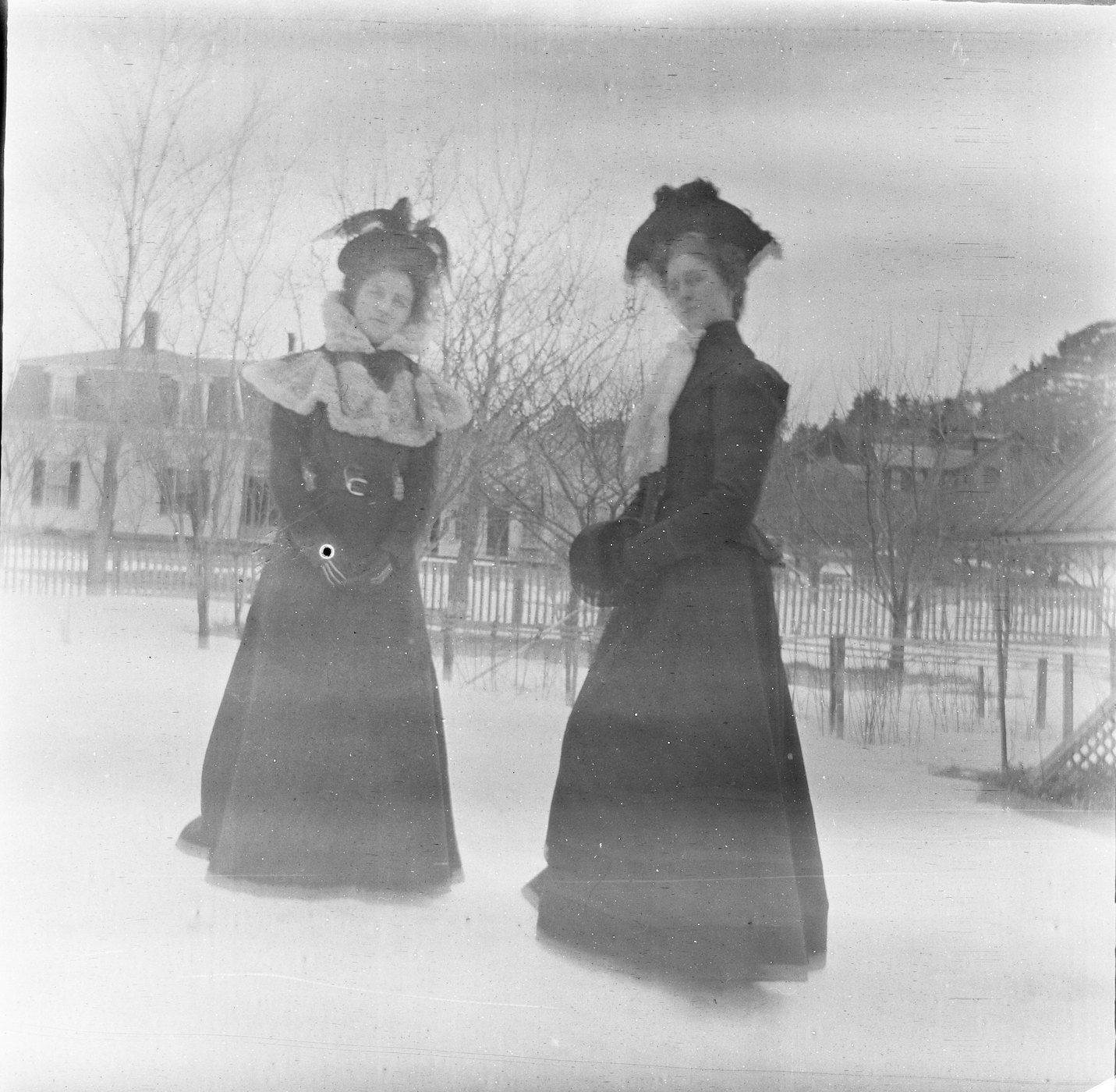 Two women in long dresses and hats stand on a snow-covered surface with buildings and trees in the background.