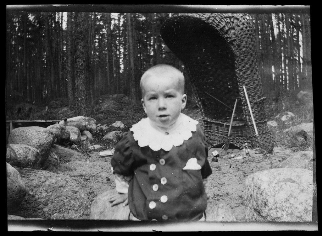 A vintage black and white photograph of a young child in a forest, standing in front of a woven tunnel.