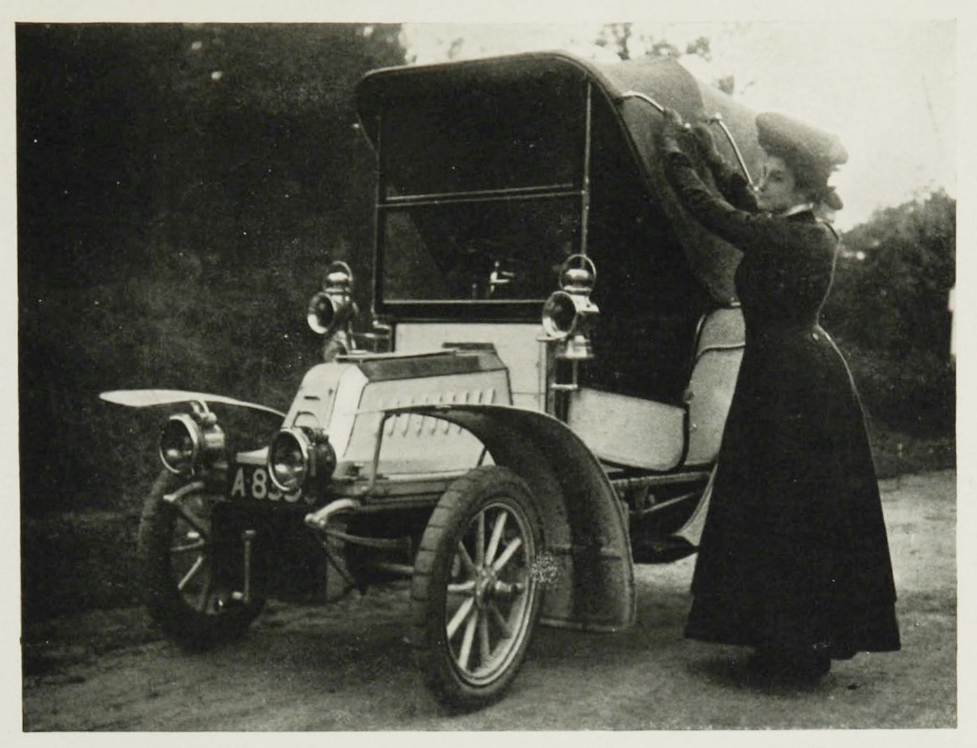 A vintage black and white photograph of a woman inspecting an early automobile.
