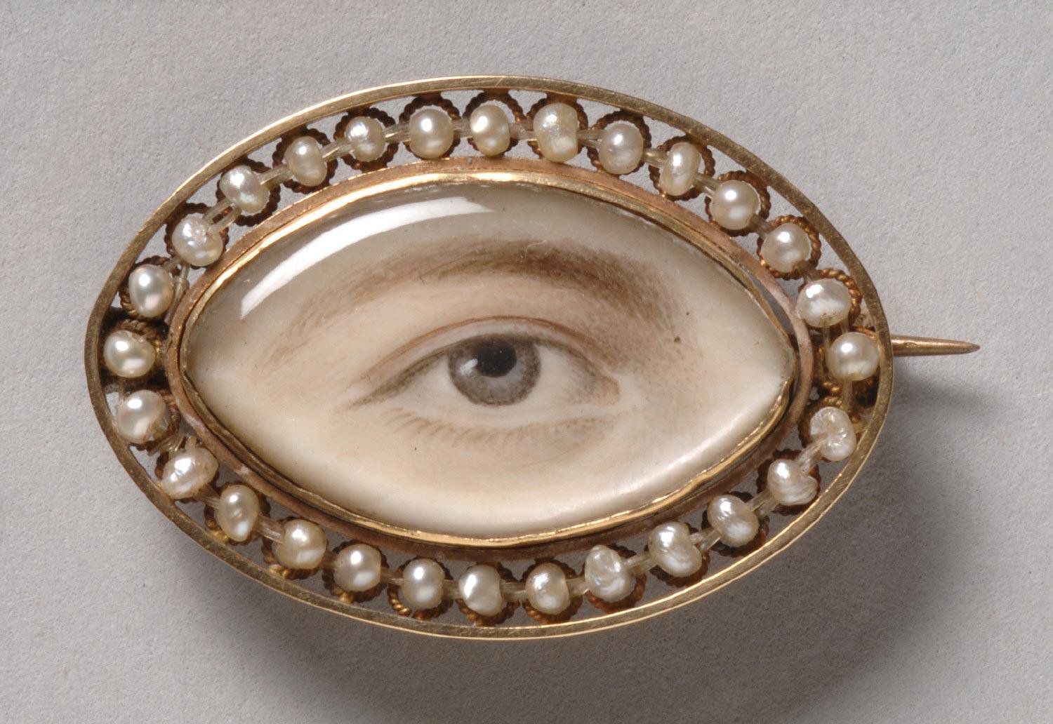 Close-up of a vintage brooch featuring a realistic painted eye surrounded by pearls and a gold frame.