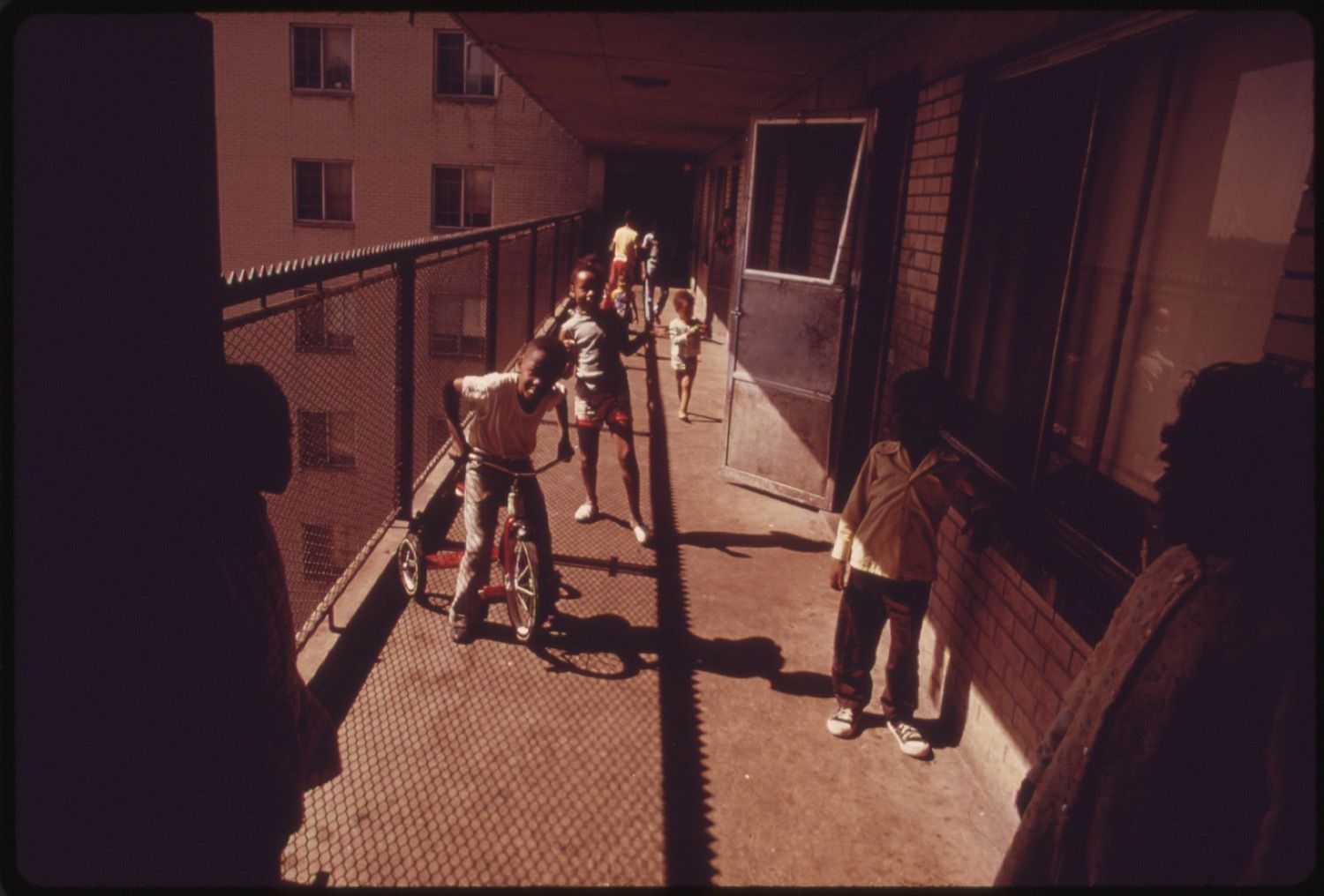 A group of children playing in a hallway of a multi-story building, with a bicycle and long shadows.