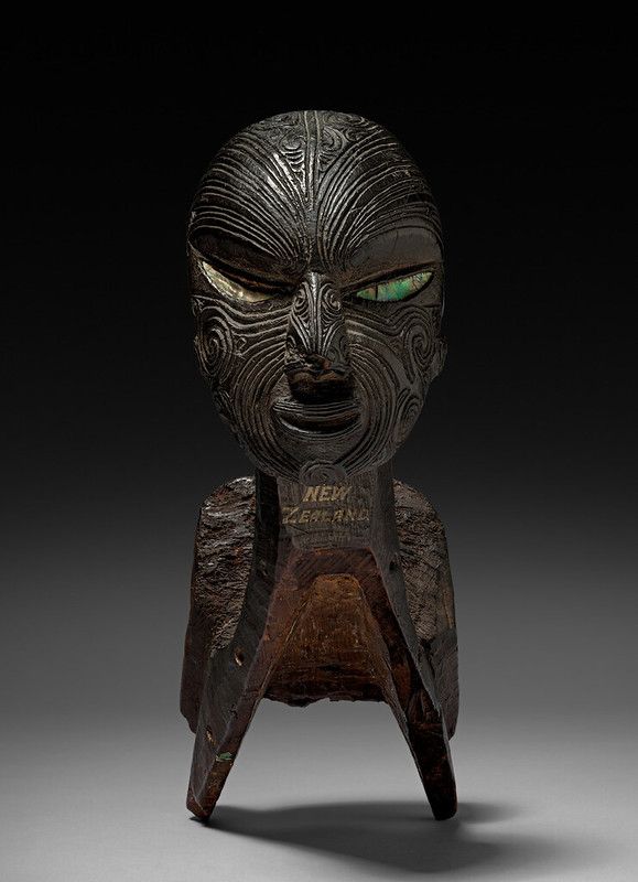 A dark wooden sculpture of a Māori head with intricate carvings and inlaid eyes, mounted on a three-pronged base.