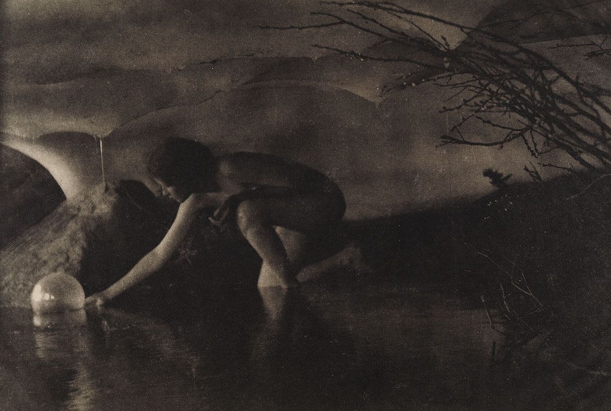 A black and white photograph of a nude woman crouching near a body of water, reaching towards it with her hand. The background features a blurred landscape with a tree and a dark sky.