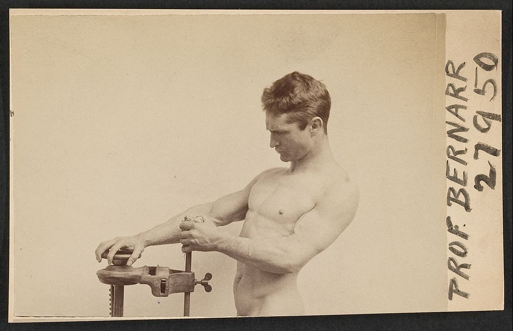 Sepia-toned photograph of a muscular man having his arm measured with a specialized machine.