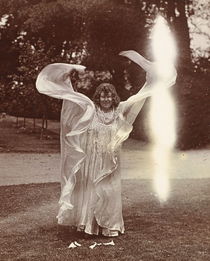 A vintage photograph of a woman in a flowing white dress with outstretched arms, draped in sheer fabric, and a bright light source behind her in a grassy outdoor setting.