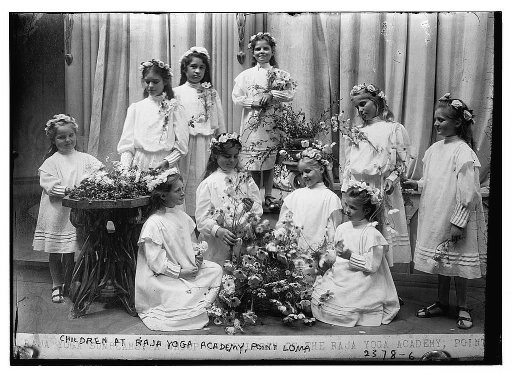 A group of children in white dresses and floral wreaths stand on a stage with flowers, likely at the Raja Yoga Academy in Point Loma, California.