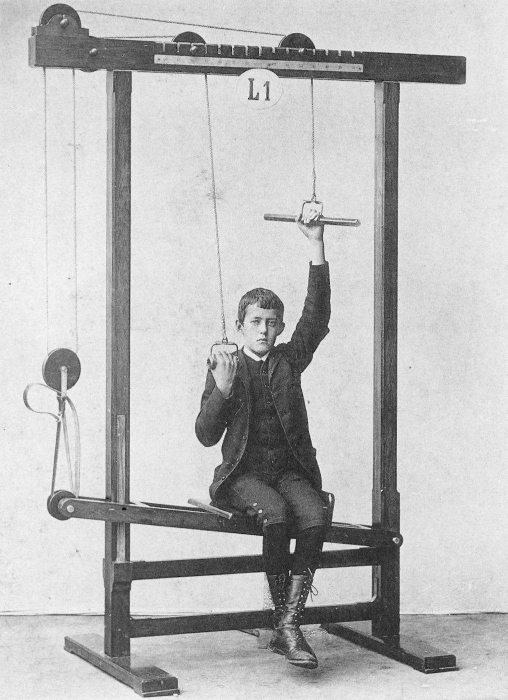 Vintage black and white photograph of a young boy using a complex exercise machine.