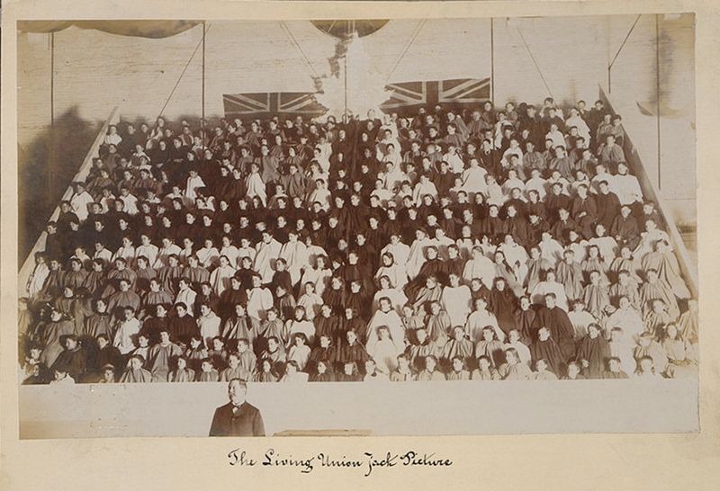 A vintage sepia-toned photograph of a large group of people arranged in a pyramidal formation, resembling a 'living picture' or tableau vivant. The group is composed of women and a single man in the foreground, all dressed in dark clothing, likely uniforms or costumes. At the very top of the pyramid, two Union Jack flags are prominently displayed. Below the image, handwritten in elegant script, is the text 'The Living Union Jack Picture'.