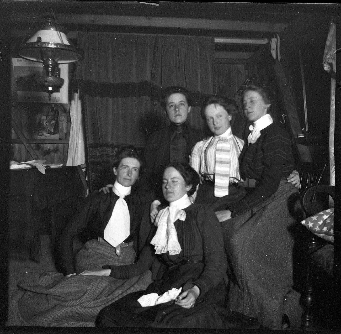 A black and white photograph of five people dressed in vintage clothing, posing indoors in front of a draped backdrop.