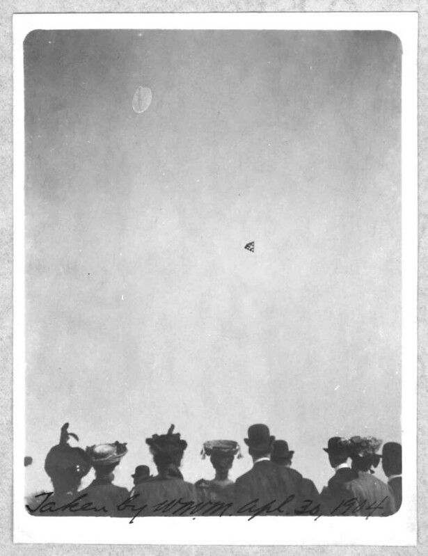 A black and white photograph of a crowd of people looking up at a biplane in the sky, dated April 23, 1914.