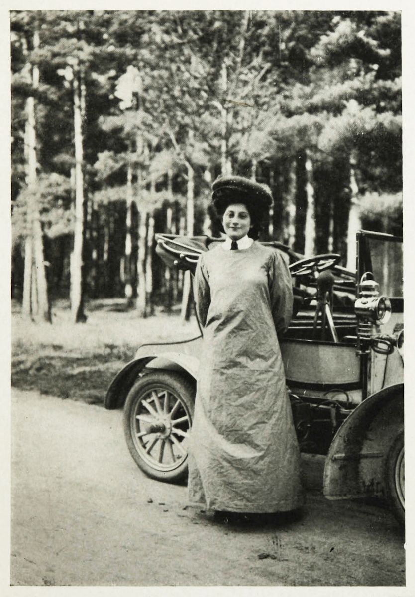 A vintage black and white photograph of a woman in a long dress and hat standing next to a vintage car in a forest.
