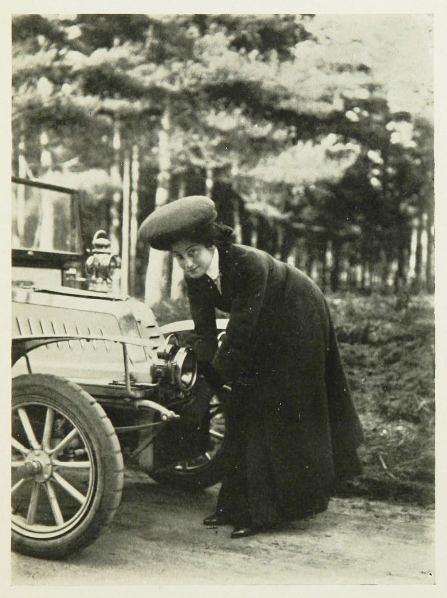 A vintage black and white photograph of a woman in a long dress and hat working on the tire of a vintage car in a wooded area.