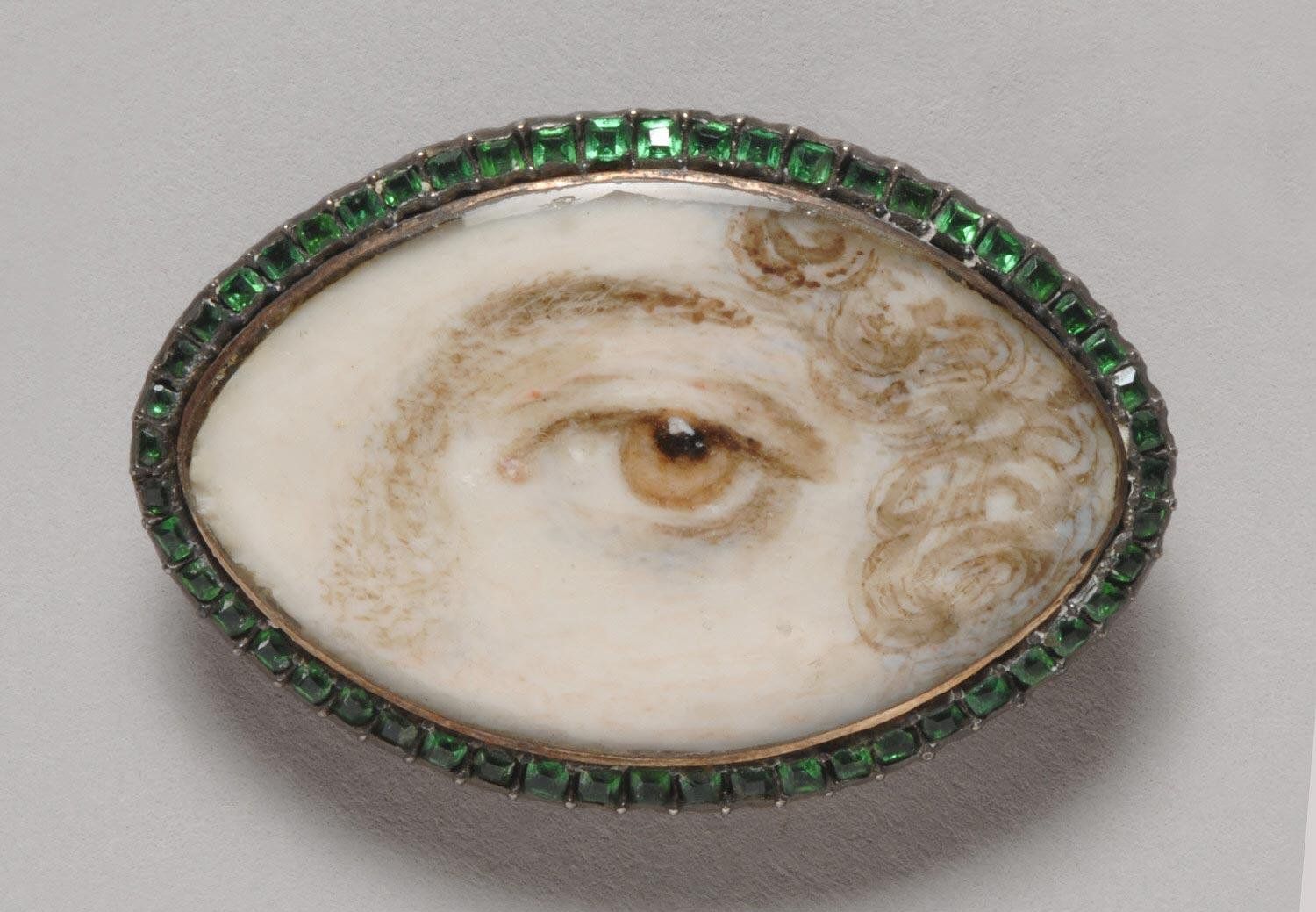 Close-up of an oval brooch with a miniature painting of a human eye on ivory, framed by a silver setting with green gemstones.