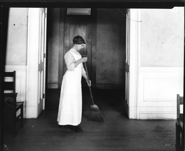 A black and white photograph of a woman in a long dress and cap sweeping the wooden floor of a church or institutional building.