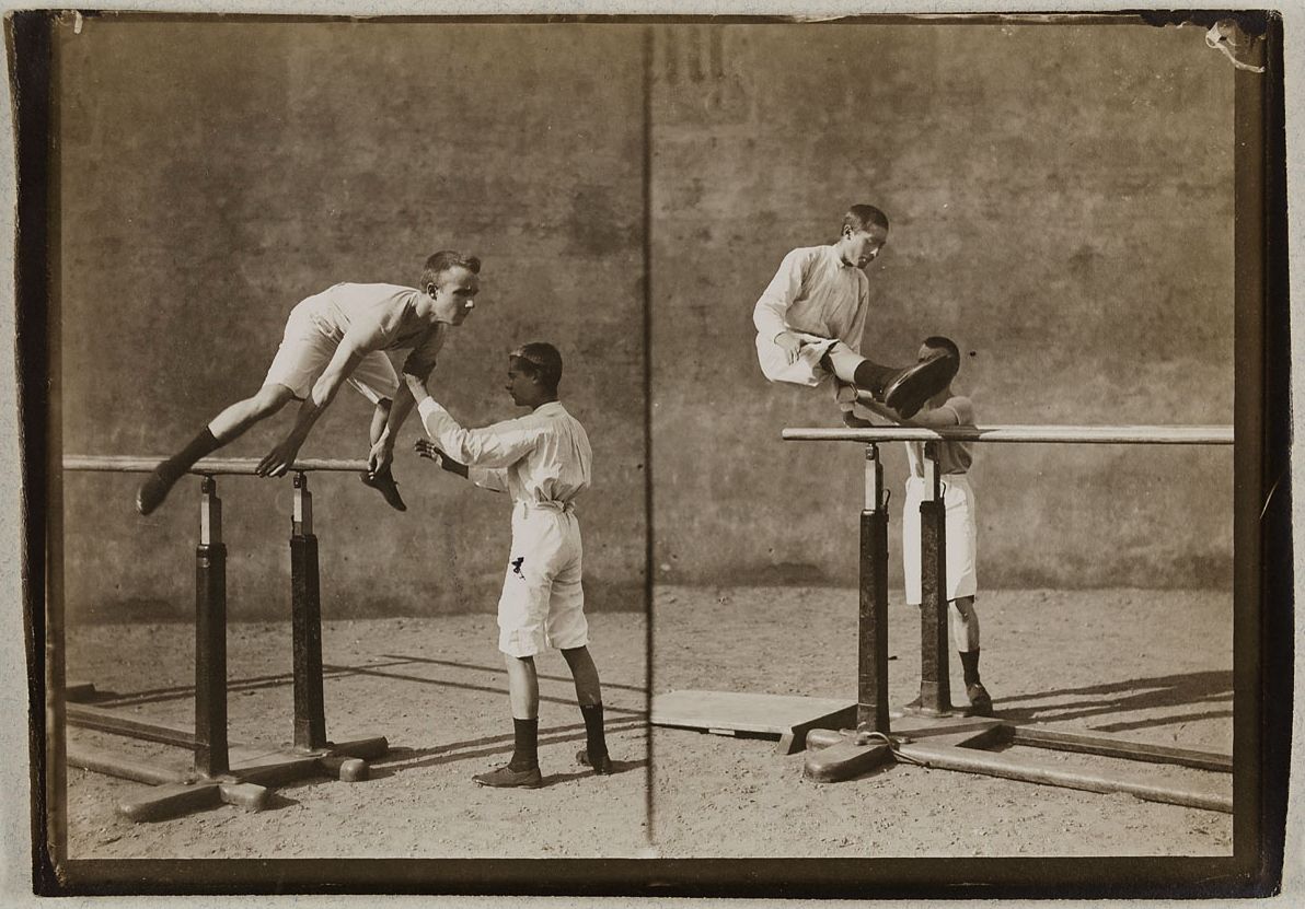 A sepia-toned photograph of a man performing a gymnastic exercise on parallel bars, with a spotter assisting him. The image is a composite, showing the gymnast in two different stages of the movement.