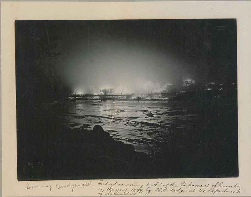 A vintage black and white photograph of a fire in Bridgwater, Canada, in 1899. The fire is centered around a dock or waterfront area, with buildings lining the shore. The fire illuminates the night sky, creating a bright, hazy glow. The water in the foreground reflects the light and smoke, creating a sense of chaos and destruction.