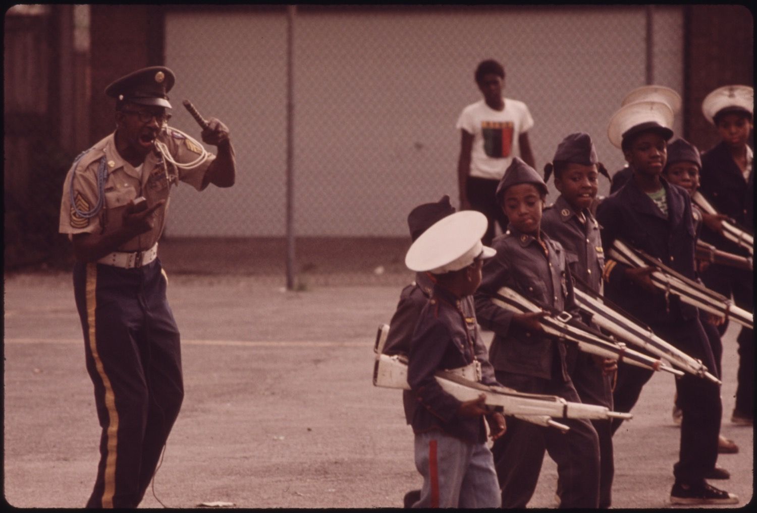 A band of young boys in uniform practicing outdoors, directed by a man in a formal band uniform.