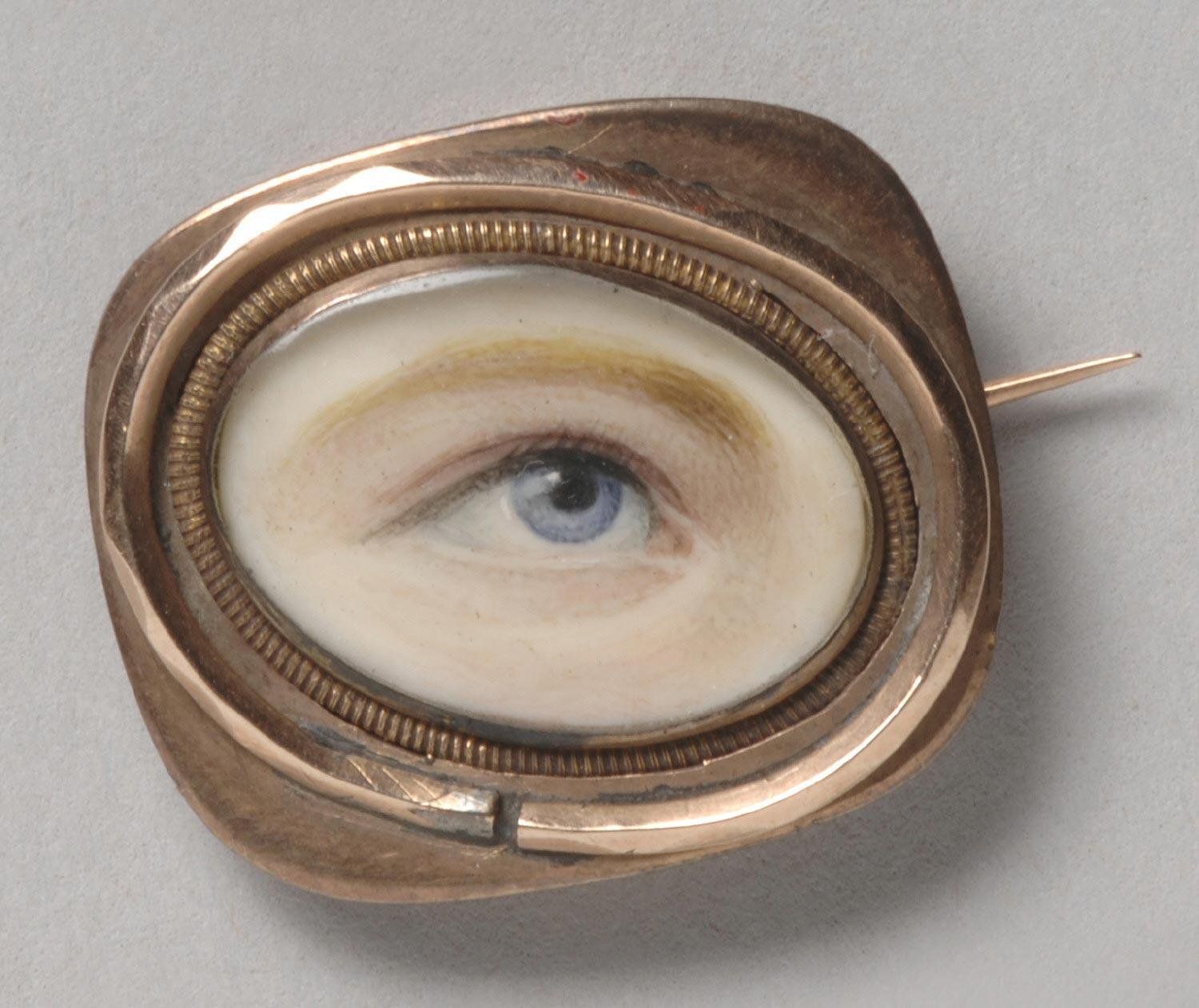Close-up of a vintage brooch featuring a realistic depiction of a human eye painted on ivory and encased in an ornate gold frame.