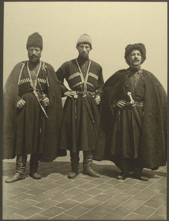A vintage sepia-toned photograph featuring three men standing in a row, dressed in traditional clothing.