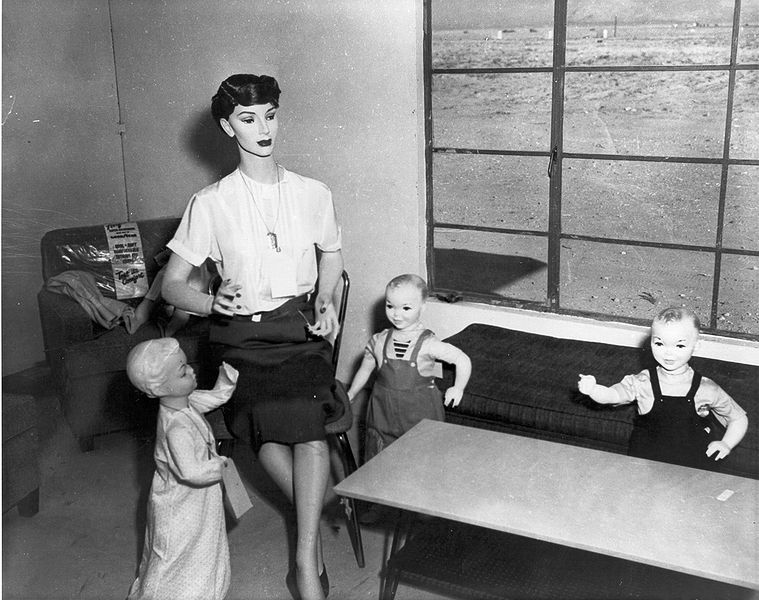 A black and white photograph of a mannequin teacher with three baby dolls as students in a vintage classroom setup.