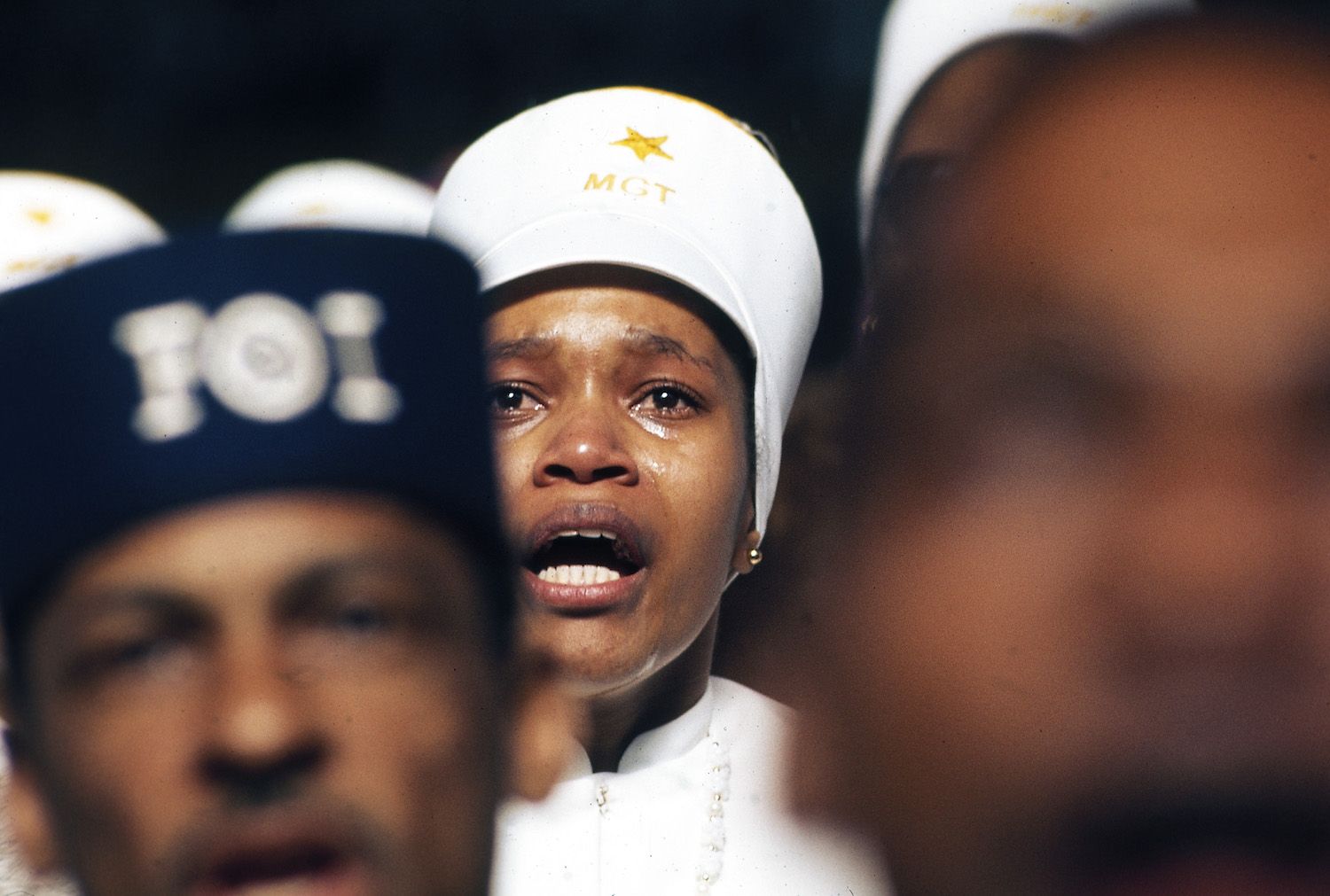 Close-up of a young Black woman wearing a white cap with 'MGT' and a star emblem, speaking with a determined expression, surrounded by others in similar caps.