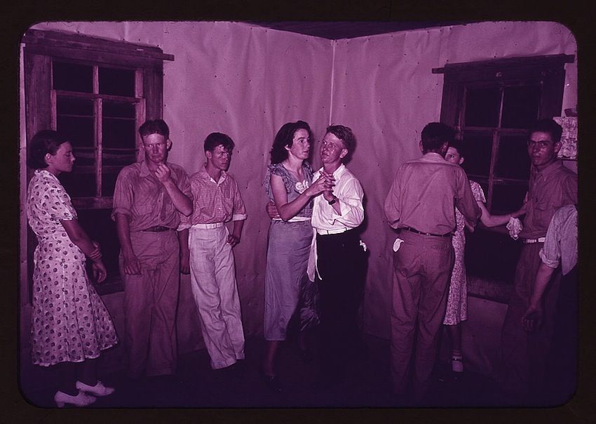 A vintage photograph of a group of young people dancing in a rustic hall. The image is dominated by a magenta hue, and the scene is filled with energy and youthful exuberance.