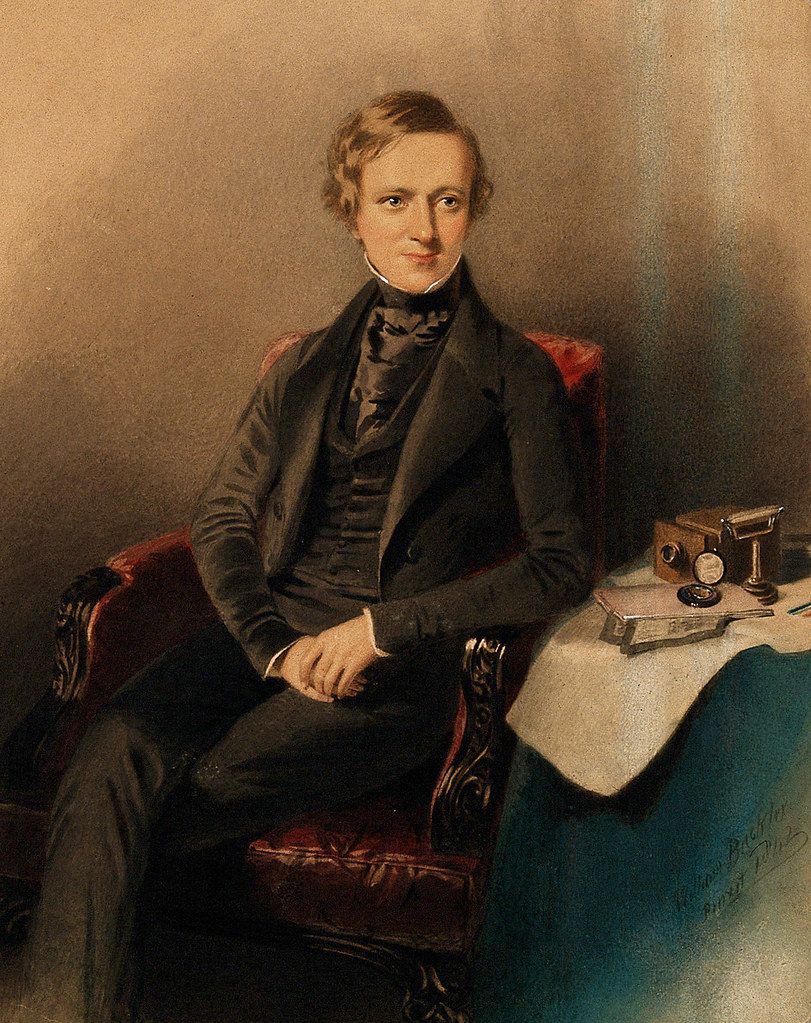 Vintage portrait of a man in formal attire seated in a red chair with scientific instruments on a table beside him.