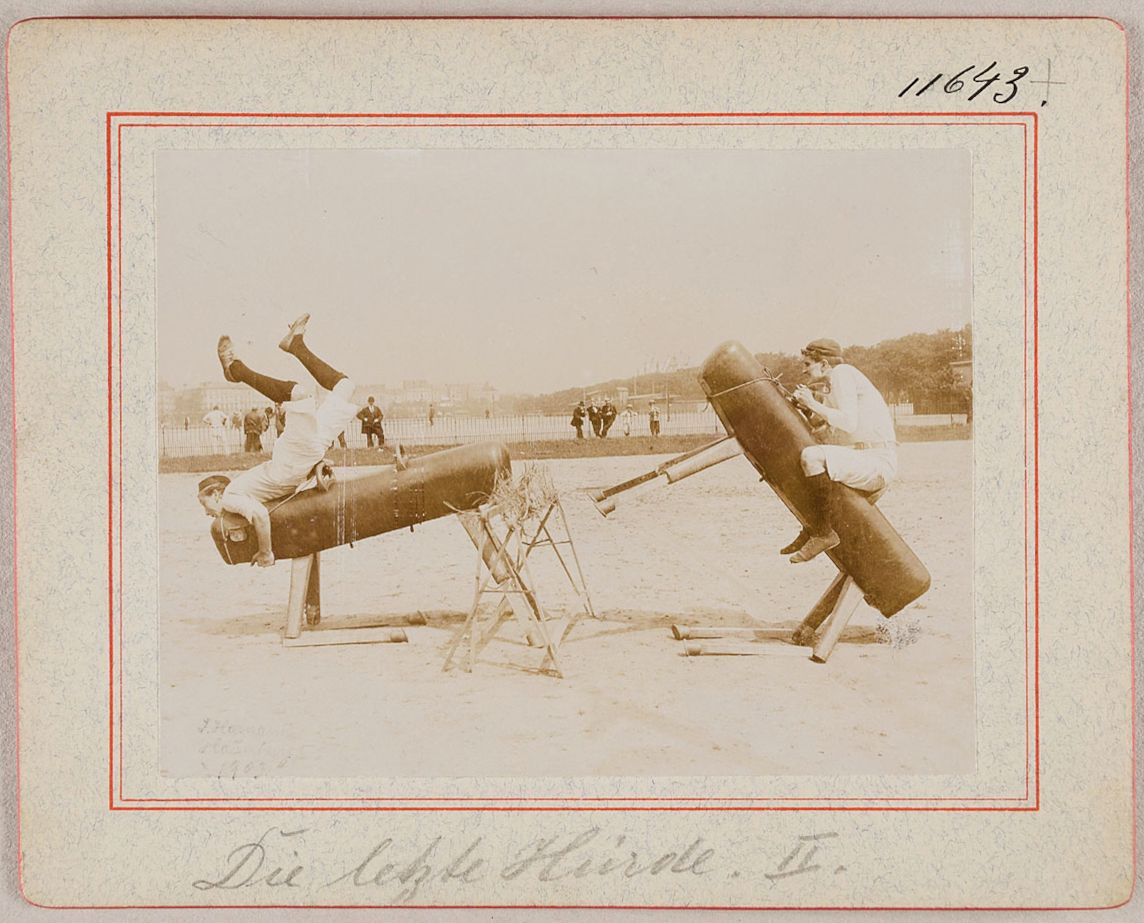 Vintage photograph of two cylindrical machines with people inside, positioned on a field with onlookers, and the text 'Die Höhle Hirde. I.' below the image.