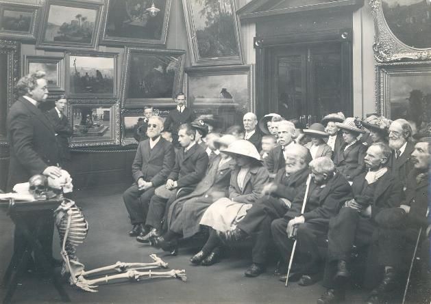 A vintage black and white photograph of a lecture with a skeleton and a group of formally dressed people.