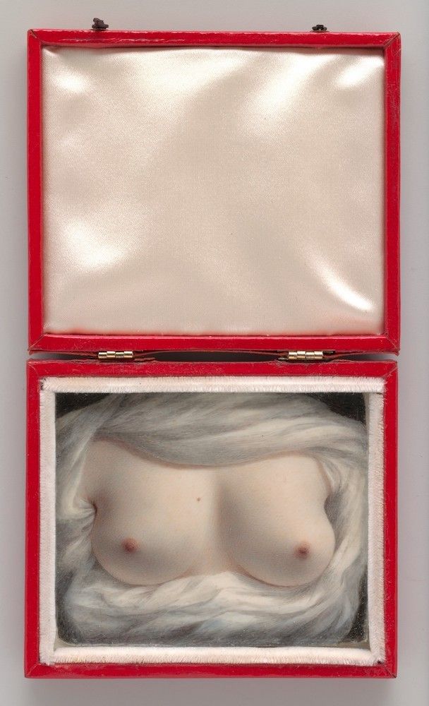 A red box with a satin lining, opened to reveal a sculpted or painted depiction of a woman's bare chest partially obscured by white fur.