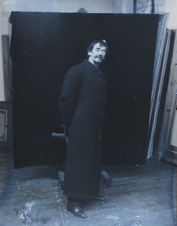 A vintage black and white photograph of a man in a long coat standing in a doorway.