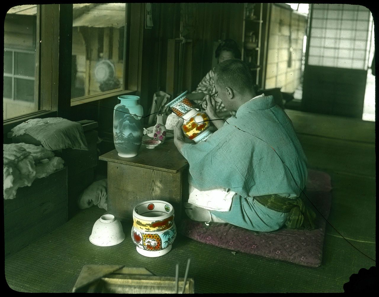 A man in a kimono is painting a ceramic vase in a traditional Japanese room.