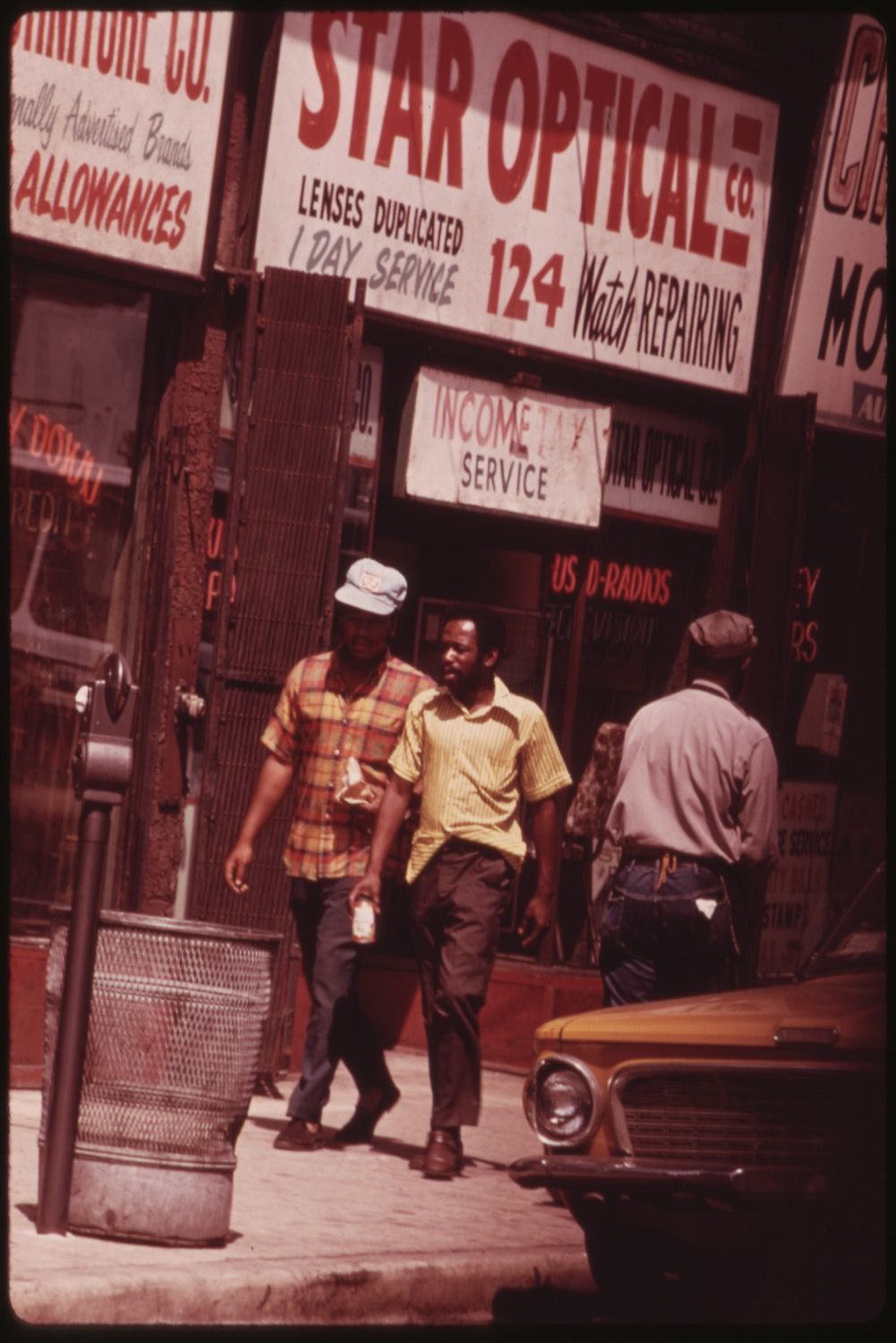 A street scene with three men walking on a sidewalk in front of a row of storefronts. The storefronts are adorned with colorful signs advertising various services, including "Star Optical Co." and "Watch Repairing."