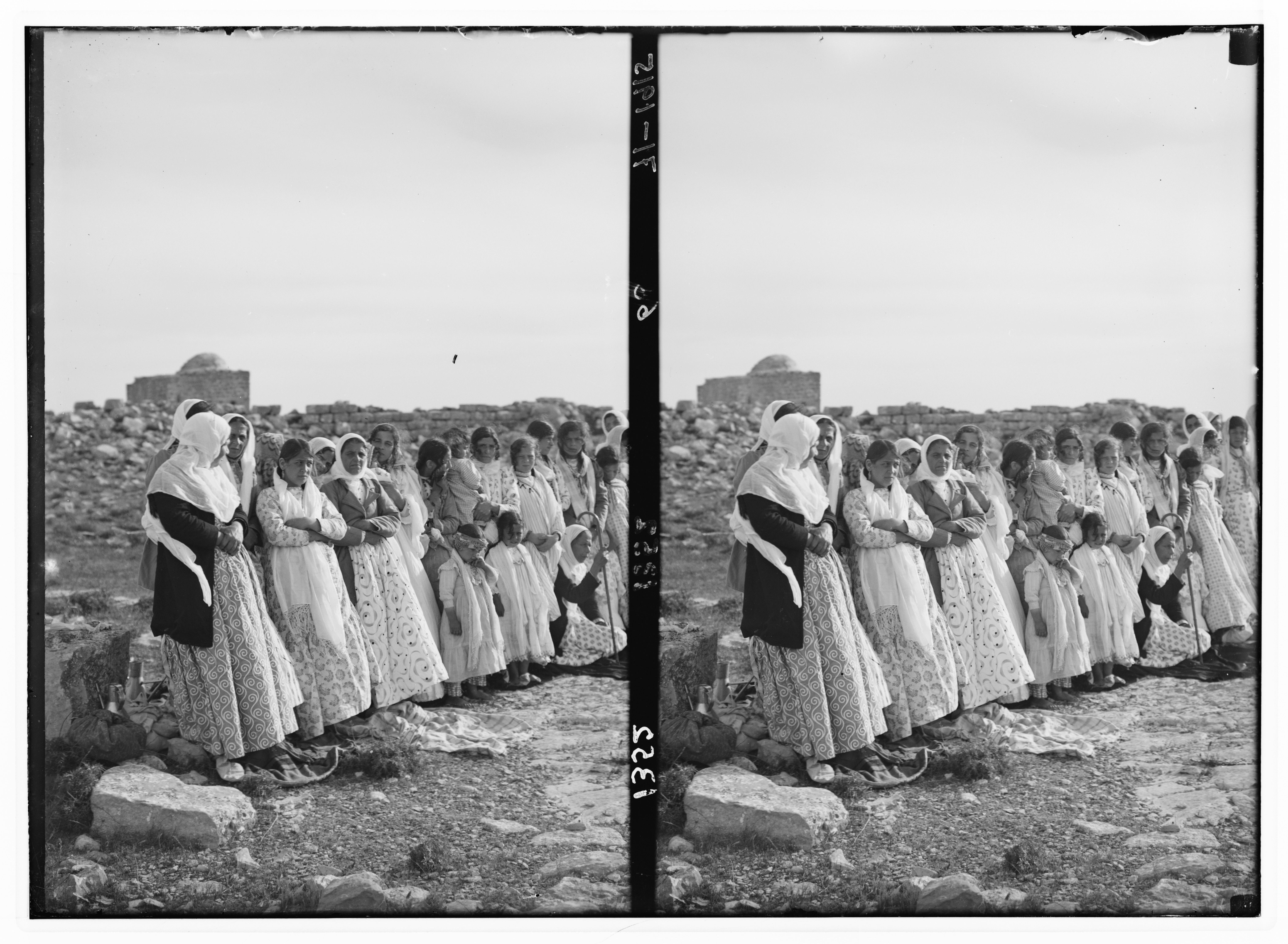 A black and white photograph of a large group of women standing in a line outdoors, dressed in long dresses and head coverings, in front of a stone wall and a building.